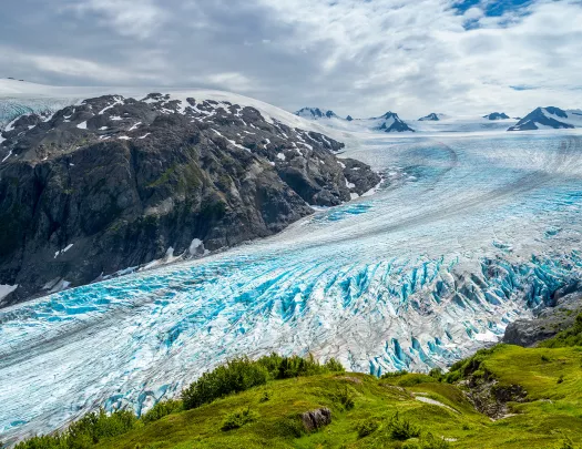 Shot of glacial vista, green cliff, blue ice, grey mountain.