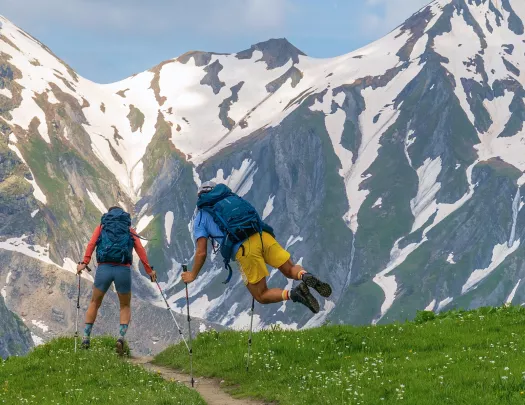 Two men using hiking poles walking on a dirt trail with snow-capped mountains in front