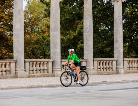 Woman wearing green biking gear, riding a bike on a road with large stone pillars in the background
