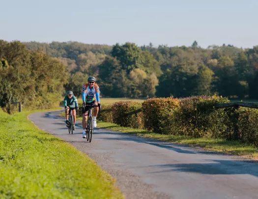 Two people smiling while riding their bikes on a road surrounded by bushes and trees