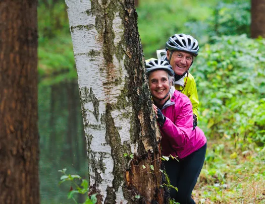 Man and woman wearing neon jackets, hiding behind a tree ina forest