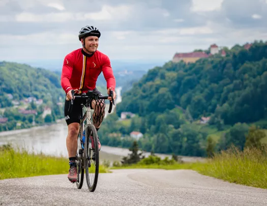 Man riding a bike on an asphalt road, with mountains of trees behind him