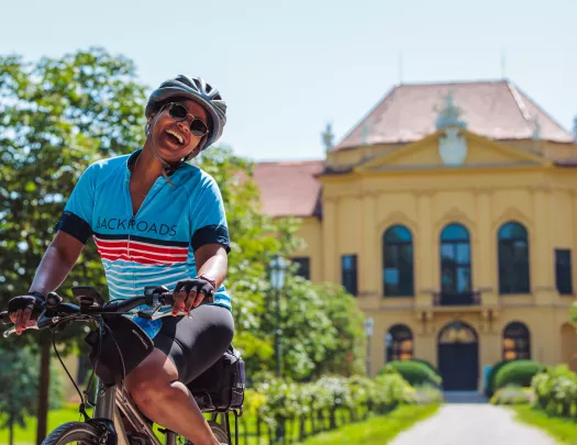 Woman wearing sunglasses and riding a bike in front of a long pathway leading to a yellow building