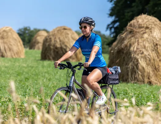 Woman biking through a grassy field with piles of hay in the background