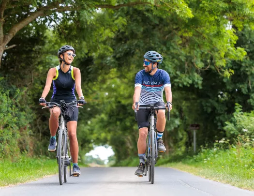 Man and woman smiling while riding their bikes on a road, with large trees surrounding