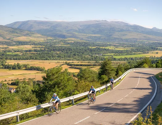 Group of 3 people riding bikes on an asphalt road, with valleys of trees in the distance