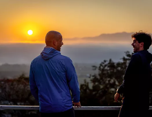Two men laughing while standing on an outdoor patio with the sunset in the background