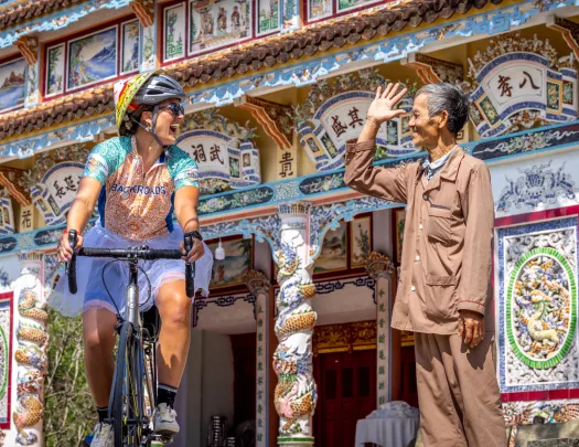 Woman riding a bike in front of a large temple, with a man smiling and waving at her