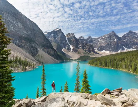 Man standing on a boulder, looking out to a blue lake and tall trees