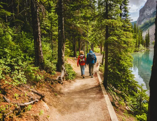 Two guests hiking down forest trail, river or lake to their right.