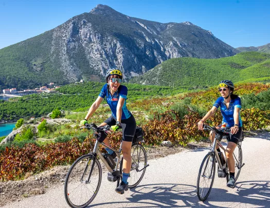 Two women smiling while riding bikes, with tall mountains in the background