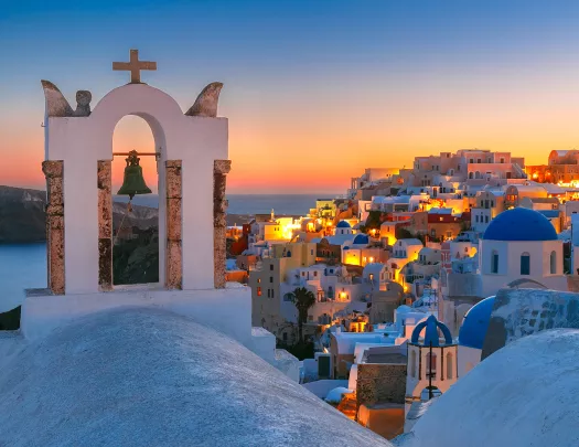 Sunset shot of Santorini, city lit up, church bell in foreground.