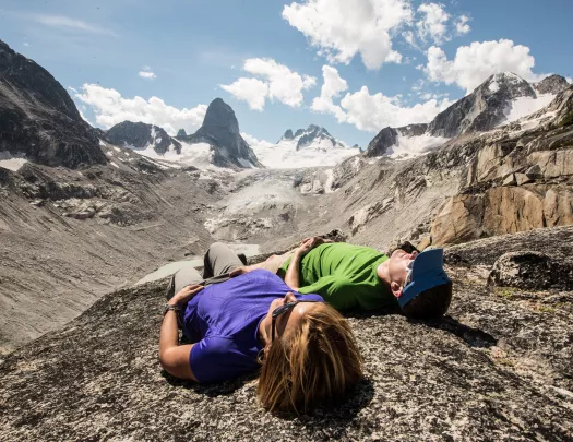 Two guests laying on rocky mountainside, arid cliffs and clouds in background.