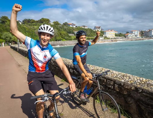 Two guests, cycling with fists raised, ocean to left.