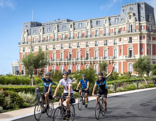 Four guests cycling past Hôtel Du Palais Biarritz, waving to camera.
