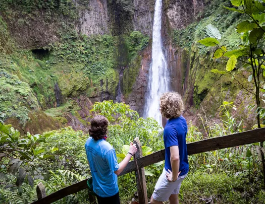 Two guests on forest path, looking down at forest, waterfall.
