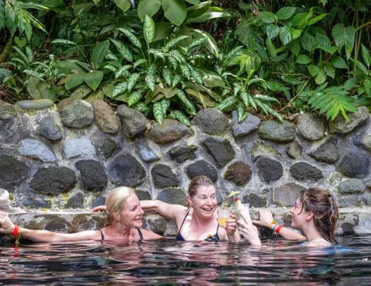 Three women holding drinks and swimming in a pool, surrounded by stone walls