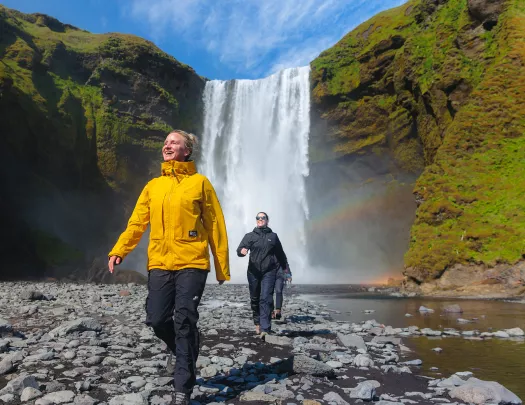 Two women smiling while walking on a rocky path in front of a waterfall