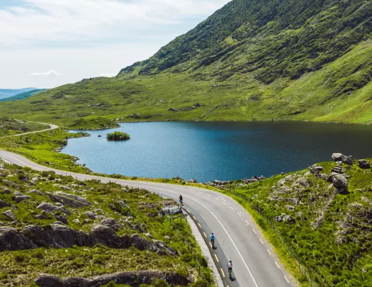 Road surrounded by large grass hills and a clear, blue lake to the right