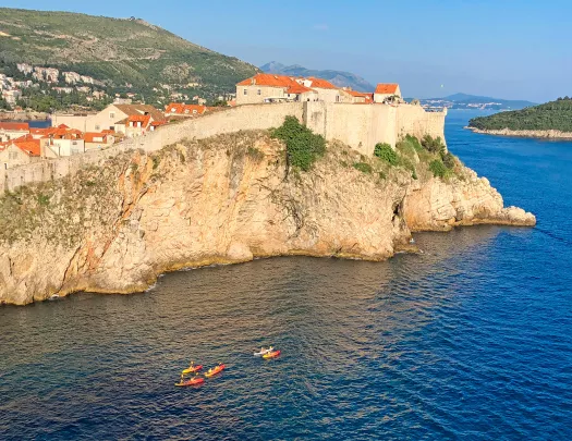 Wide shot of walled cliffside houses, guests kayaking below.