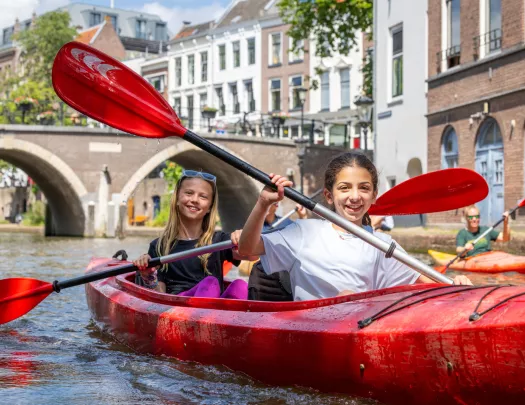 Two girls paddling in a red kayak on a river, with a town in the background