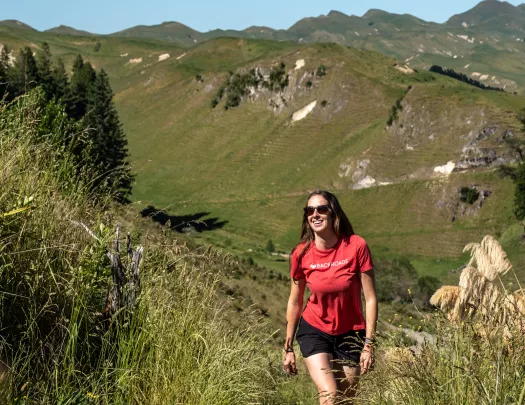 Woman wearing a red shirt, smiling while walking up a grassy trail on a hill