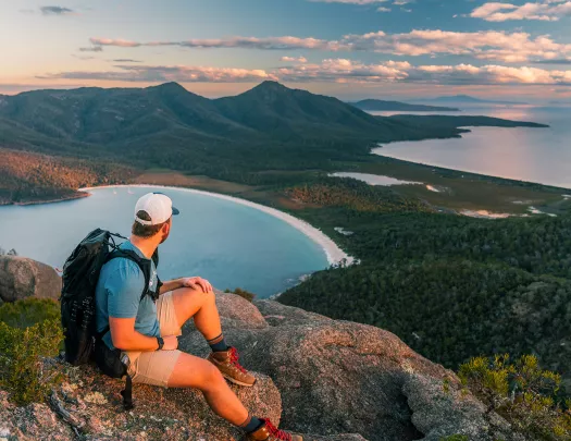 Man sitting on a cliff looking out to a valley of trees and a lake
