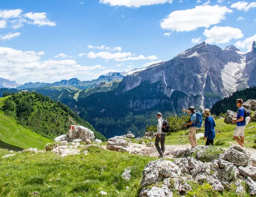 Group of guests hiking down mountain trail, Dolomite range in background.