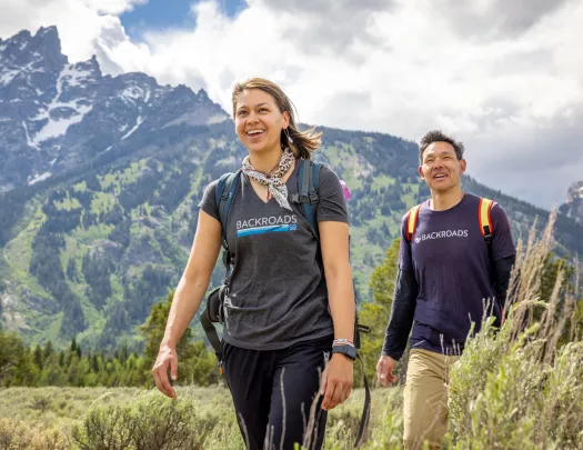 Man and woman smiling while walking through a grassy valley, with large mountains in the background