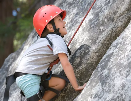 Boy wearing a helmet, attached to a harness climbing up a boulder