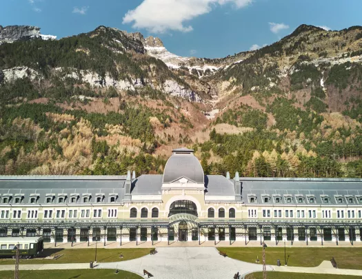 Wide tan and gray building with a large, gray courtyard and tall mountains in the background