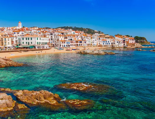 Shot of Costa Brava coastline, blue water, tan and white buildings.