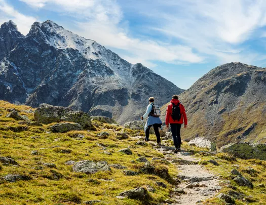 Man and woman ascending a rocky, dirt trail with large mountains ahead