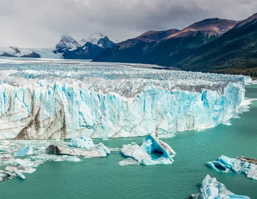Wide shot of large glacial shelf, dark clouds and mountains in background.