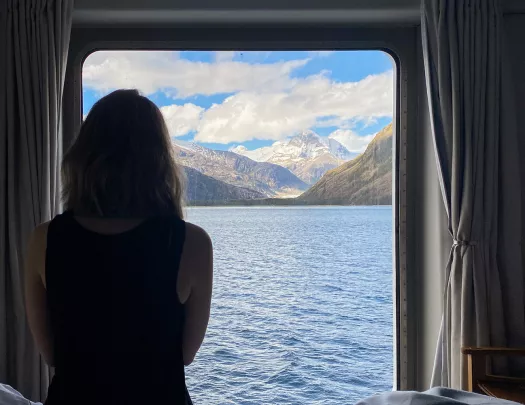 Guest on cruise ship in room, looking out towards mountains.