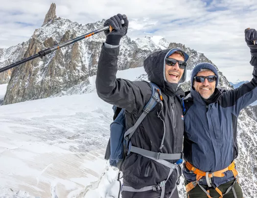 Two men in snow gear, holding up their arms while holding walking poles on top of a snowy mountain