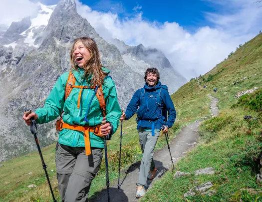 Man and woman with walking poles, hiking on a dirt trail with large mountains in the background
