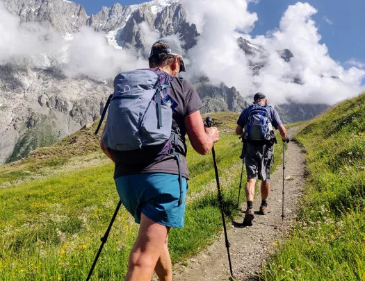 Two guests walking up mountain trail, clouds, mountain in distance. 