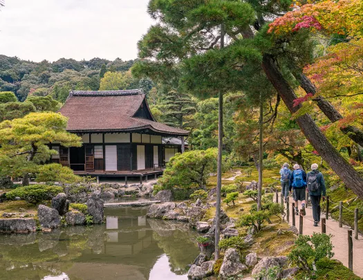 Three people walking on a small path on the right towards a Japanese-style building in the forest