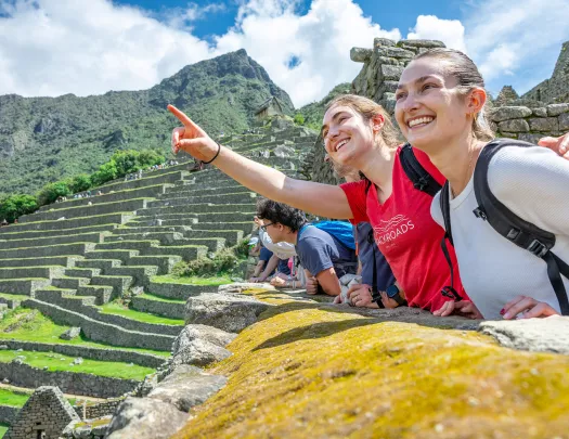 Two women smiling and pointing towards the sky, with large stone ruins in the background