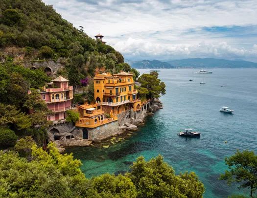 Wide shot of coastal town, two buildings on hill, one red, one gold. Ships in water below.