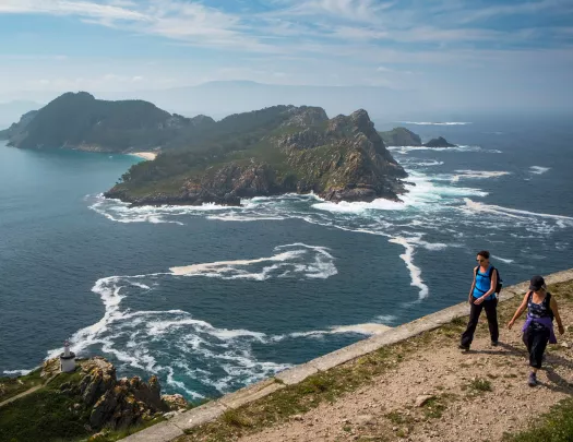 Two guests on sheer cliffside, large craggy island, ocean behind them.